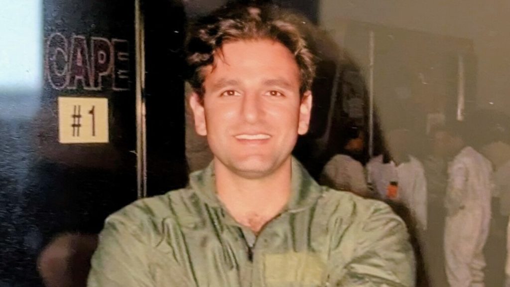 Man in a green jumpsuit smiling with "CAPE #1" sign in the background.