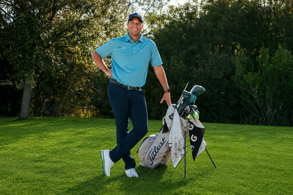 A golfer in a blue shirt leans on a golf bag, smiling, with trees in the background.