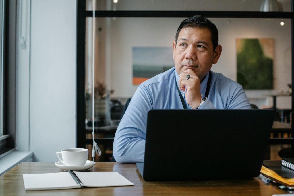 Man in a blue shirt sitting at a desk with a laptop, looking thoughtful.