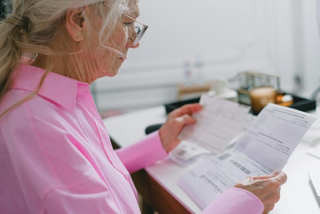 An elderly person in a pink shirt examines documents at a desk.
