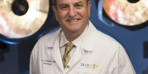Smiling man in a white lab coat stands in an operating room with overhead lights.