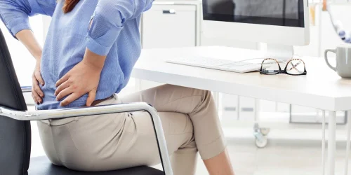Woman holding her back in pain while sitting at an office desk.