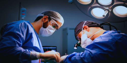 Two surgeons in masks and scrubs perform surgery under operating room lights.