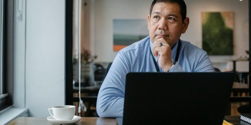 Man in a blue shirt sitting at a desk with a laptop, looking thoughtful.