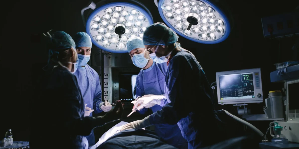 Surgeons in blue scrubs perform surgery under bright lights in an operating room.